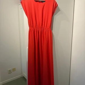 Lång klänning  - En väldigt fin klänning som har en unik rosa färg. Färgen har en lite rödaktig nyans/underton och är väldigt somrig. Den är köpt på Vero Moda och har använts 1 gång. Den har även ett band som man kan knyta klänningen med och är väldigt söt inför sommaren, rekommenderar starkt! Säljer den då den inte längre passar💗 (Kameran visar en mer rödaktig färg, dock ser den inte ut som den gör på kameran utan den har en mer rosa färg i verkligheten)