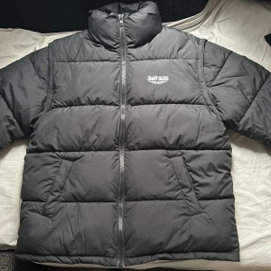 Svart pufferjacka från Sweet SKTBS - Säljer en svart pufferjacka från Sweet SKTBS. Jackan har en dragkedja framtill och är perfekt för kyligare väder. Den är designad med en hög krage för extra värme och stil. Märkets logga är tryckt på bröstet. En riktigt schysst jacka för vintern! samt en schysst väst när man känner för det, så det är dragkedja vid armhålen för att kunna ta av och på armarna. den är större än man tror…