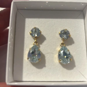 Guldörhängen med blå stenar - Eleganta örhängen i guld med två blå stenar i varje örhänge. Den övre stenen är rund och den nedre är droppformad, vilket ger en stilfull look. Perfekta för att lägga till en touch av glamour.