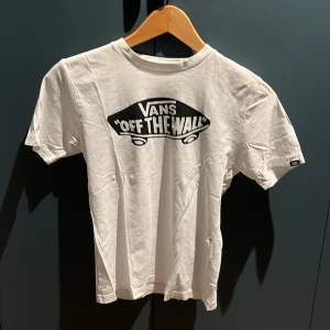Vit t-shirt från Vans - Säljer en klassisk vit t-shirt från Vans med det ikoniska 'Off The Wall'-trycket i svart på framsidan. T-shirten har en normal passform och är tillverkad i bomull, vilket gör den bekväm och lätt att bära.Barn storlek på denna