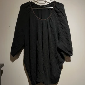 Svart blus med hals detalj - Snygg svart blus med en unik detalj vid halsringningen. Blusen har trekvartsärmar och ett löst fall som ger en avslappnad look. Perfekt för att lägga till en touch av elegans i din garderob.