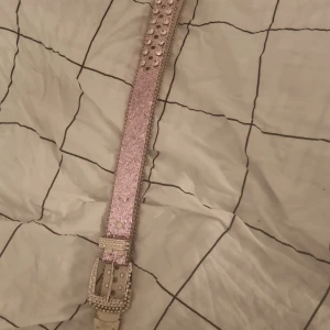 Glittrigt rosa bälte med nitar - Säljer ett glittrigt rosa bälte dekorerat med silverfärgade nitar och strassdetaljer. Bältet har en rektangulär spänne med liknande dekorationer. Perfekt för att ge en extra touch till din outfit!