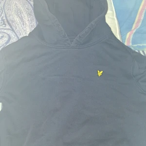 hoodie från lyle & scott - Säljer en stilren svart hoodie från Lyle & Scott med deras ikoniska gula logga på bröstet. Perfekt för en avslappnad look. Den har en klassisk känguruficka och en bekväm huva.