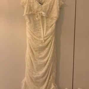 Vit spetsklänning från NLY One - Säljer en vit spetsklänning från NLY One. Klänningen har en vacker volangdetalj vid axlarna och en knytning framtill. Den är i en tight passform och man kan välja hur lång eller kort man vill ha den genom att justera banden i sidan