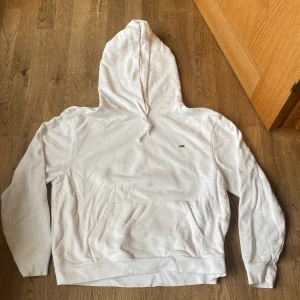 Vit hoodie från Tommy Hilfiger - Säljer en stilren vit hoodie från Tommy Hilfiger med klassisk logga på bröstet och TJ-broderi på ärmen. Hoodien har en stor ficka framtill och justerbar huva med dragsko. Perfekt för en avslappnad look.