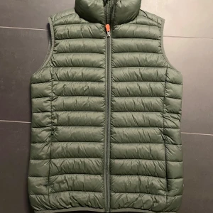 Grön dunväst från Save The Duck - Säljer en grön dunväst från Save The Duck. Västen är quiltad och har en dragkedja . Perfekt för att hålla värmen under kyliga dagar. Helt oanvänd.