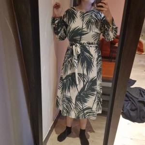 Vit och grön klänning med palmbladsmönster - Snygg vit klänning med grönt palmbladsmönster och knytskärp i midjan. Klänningen har långa ärmar och når till mitten av vaden. Perfekt för en somrig look! 🌿