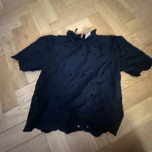 Svart blus från Holly & Whyte - Säljer en svart blus från Holly & Whyte med korta ärmar och dekorativa hål. Perfekt för en stilren look. Blusen är tillverkad i ett lätt material och har en elegant design.