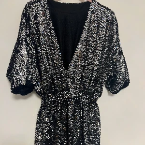 Svart glittrig kort  klänning - Snygg svart klänning täckt med glittriga paljetter. Den har en v-ringad design och korta ärmar, perfekt för en festlig kväll. Klänningen har en elastisk midja som ger en smickrande passform.