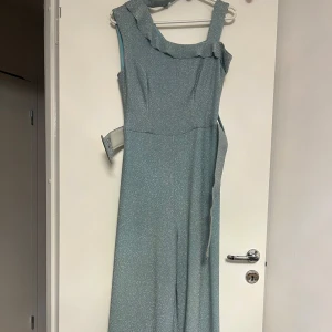 Glittrig ljusblå heldräkt  - Säljer en elegant ljusblå heldräkt med glittrigt material och volangdetaljer vid axlarna. har en smickrande passform och är perfekt för speciella tillfällen. Den är ärmlös och har en vacker fallande design. Perfekt för att glänsa på festen! ✨