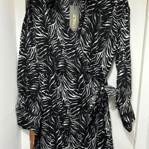 Svart och vit klänning från Only XL i storleken  - Snygg svart och vit klänning från Only med zebramönster. Klänningen har långa ärmar och en omlott-design med knytning i midjan. Perfekt för att sticka ut med stil!
