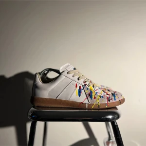 Maison margiela  - Unika sneakers med en vit bas och färgglatt stänkmönster i blått, rött, gult och svart. Skorna har en klassisk snörning och en brun sula. Perfekta för dig som vill sticka ut med en konstnärlig touch.