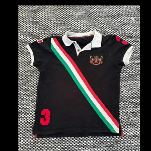 Svart kortärmad pikétröja från Adrian Hammond - Snygg svart pikétröja med vita detaljer och en diagonal rand i grönt, vitt och rött.