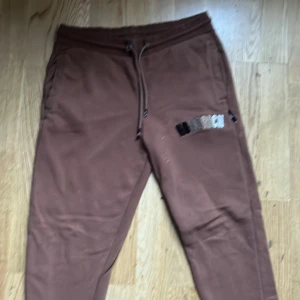 Bruna mjukisbyxor från Hoodrich - Säljer ett par bruna mjukisbyxor från Hoodrich med broderad logga på benet. Byxorna har snörning i midjan för justerbar passform och är perfekta för en avslappnad stil.