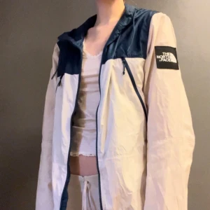 Vit och marinblå vindjacka från The North Face - Säljer en stilren vit och marinblå vindjacka från The North Face. Jackan har en dragkedja framtill och en justerbar huva. Perfekt för blåsiga dagar och har en klassisk passform.