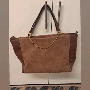 Brun axelväska med kedjedetalj - Snygg brun axelväska med en elegant kedjedetalj framtill. Väskan har både handtag och en justerbar axelrem! Använt ett par gånger 