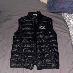 Armani EA7 väst  - Snygg svart dunväst från EA7 med dragkedja framtill. Perfekt för lager-på-lager under kyligare dagar. Västen har en stilren design med EA7-logga på bröstet.