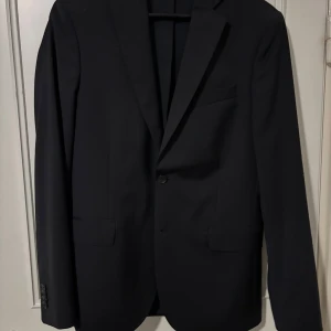 Svart kavaj från J.Lindeberg - Elegant svart kavaj från J.Lindeberg med klassisk design. Kavajen har en enkelknäppning och är tillverkad i ett stilrent material. Perfekt för formella tillfällen. Med ett par kostymbyxor från dressman.   Ny pris för hela kostymen 3600kr.                                  Pris kan diskuteras 