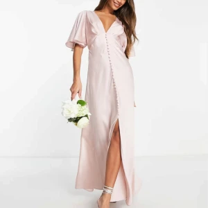 Ljusrosa långklänning med v-ringning -  Ljusrosa långklänning i satin med v-ringning och kortärmade vida ärmar. Klänningen har en slits framtill och knappar längs med framsidan. Perfekt för bal,lärarmiddagen, speciella tillfällen osv. Aldrig använd med prislapp kvar💕Nypris: 1069kr. Skriv för egna bilder💖