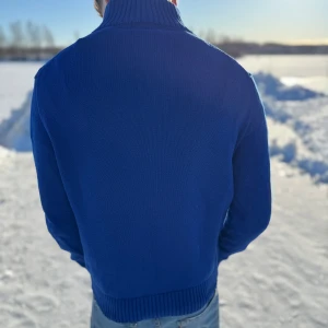 Blå stickad tröja från Ralph Lauren - Säljer en snygg blå stickad tröja från Ralph Lauren med dragkedja vid halsen och det klassiska röda logotypbroderiet på bröstet. Tröjan har långa ärmar och ribbade muddar för en bekväm passform. Perfekt för kyliga dagar!