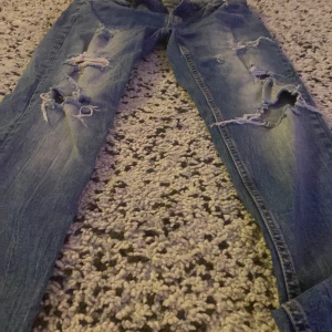 Blå slitna jeans från New Yorker - Säljer ett par blå jeans från New Yorker i bra skick. De har en cool, sliten look med flera revor och slitningar. Perfekta för en avslappnad stil! Tillverkade i 98% bomull och 2% elastan för en bekväm passform.