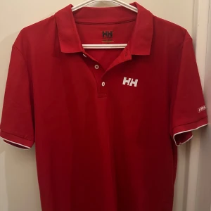 Röd pikétröja från Helly Hansen - Säljer en snygg röd pikétröja från Helly Hansen i storlek L. Perfekt för både vardag och sportiga aktiviteter. Den har coola detaljer som 'FASTER' på ärmen och ett tryck med segelbåtar och 'HH 1877' på sidan. Perfekt för dig eller en present till en kompis eller någon i familjen. Aldrig använd!!
