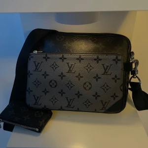 Axelväska med monogrammönster - Snygg axelväska i svart och grått med klassiskt monogrammönster. Väskan har en justerbar axelrem och en dragkedja för säker förvaring. Perfekt för både vardag och festliga tillfällen. Den är i mycket bra skick och redo att användas direkt!