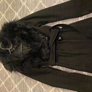 kappa  - Säljer en elegant svart kappa i storlek 34 med mjuk pälsdetalj runt kragen. Den har knappar framtill och ett bälte i midjan för en snygg passform. 