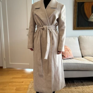 Beige kappa från NA-KD - Säljer en superfin beige kappa från NA-KD i storlek 38. Den har en klassisk design med knytskärp i midjan och långa ärmar. Perfekt för höst och vår! Kappan är i mycket bra skick och passar både till vardags och fest. 🧥✨