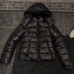 Svart dunjacka med huva - Säljer en svart dunjacka i mycket bra skick. Den har en praktisk huva och dragkedja framtill. Perfekt för kalla vinterdagar. Jackan är långärmad och har en stilren design som passar till det mesta. Håll dig varm och snygg i vinter!