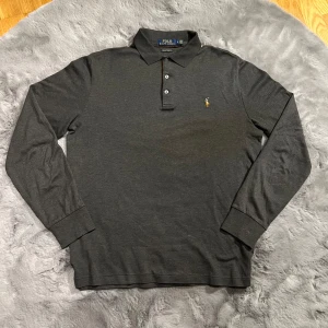 Grå tröja från Ralph Lauren - Säljer en stilren grå tröja från Ralph Lauren i storlek M. Tröjan har långa ärmar och en klassisk krage med tre knappar. Den är i ett snyggt slim fit och har det ikoniska broderade logotypen på bröstet. Perfekt för både vardag och fest!