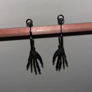 Svarta skelettörhängen - Coola svarta örhängen i form av skelett händer. Perfekta för en unik och edgy stil, kanske till Halloween eller en rockig fest. De hänger från små svarta ringar och ger en riktigt cool look. Passar perfekt för dig som vill sticka ut!