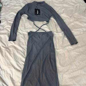 Grå croppad klänning med omlott - Snygg grå croppad klänning med långärmad topp och omlottkjol. Perfekt för en stilren look. Materialet är mjukt och stretchigt, vilket ger en bekväm passform. Den har en unik design med snörning i midjan.