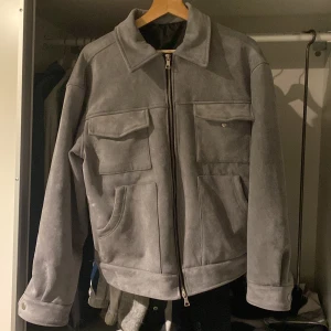 Grå mockajacka - Säljer en snygg grå mockajacka i mycket bra skick. Jackan har en dragkedja framtill och två stora fickor med lock. Perfekt för höst och vår, med en stilren och tidlös design. Passar både till vardags och lite finare tillfällen.