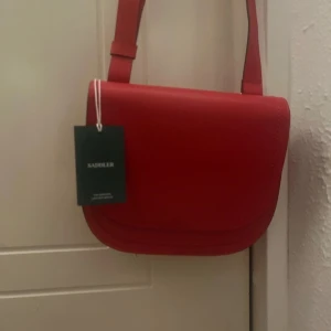 Röd axelväska från Saddler - Snygg röd axelväska från Saddler i skinn. Perfekt för både vardag och fest. Väskan har en justerbar axelrem och ett rymligt fack med dragkedja inuti. Den är i nytt skick med etikett kvar. En stilren och praktisk accessoar för alla tillfällen!