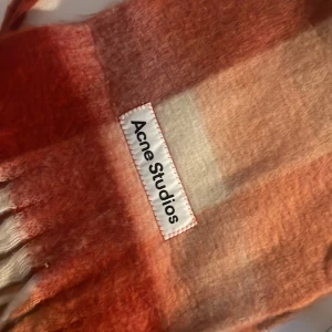 Rutig halsduk från Acne Studios - Säljer en superfin rutig halsduk från Acne Studios i varma toner av röd och beige. Den är gjord i ett mjukt material som känns lyxigt och perfekt för kyliga dagar. Märkeslappen är snyggt fastsydd med röd tråd. Perfekt accessoar för att hålla stilen i höst och vinter! 🍂❄️