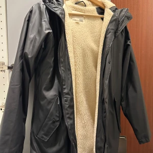 Svart regnjacka med teddyfoder - Säljer en svart regnjacka från 157 med mysigt beige teddyfoder. Perfekt för regniga dagar med sin långa design och praktiska huva. Jackan har dragkedja och knappar framtill för extra skydd mot väder och vind. Den är i mycket bra skick och passar perfekt för höst och vår.
