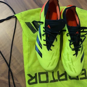 Adidas Predator fotbollsskor - Säljer ett par grymma Adidas Predator fotbollsskor i neonfärg med svarta och röda detaljer. Skorna har snörning och är perfekta för fotbollsträning eller match. De är i mycket bra skick och kommer med en praktisk förvaringspåse. Perfekt för dig som vill sticka ut på planen!