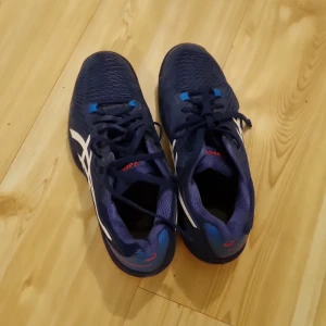 Blå Asics tennisskor - Säljer ett par blå Asics tennisskor i bra skick. De har en snygg design med vita detaljer och snörning. Perfekta för träning eller vardagsbruk. Storlek 40,5. Skorna är bekväma och ger bra stöd för fötterna.