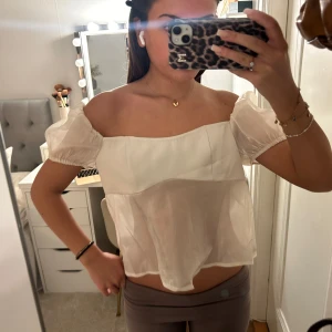 Vit offshoulder blus - Säljer en söt vit offshoulder blus med puffiga korta ärmar och öppen rygg med knytband. Perfekt för sommardagar eller en festlig kväll. Blusen är luftig och har en romantisk stil.