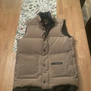 Beige dunväst från Canada Goose - Säljer en snygg och varm beige dunväst från Canada Goose i storlek XXS. Perfekt för kyliga dagar med sin quiltade design och praktiska knappar. Västen är i mycket bra skick och passar perfekt för höst och vinter. Ett måste för garderoben! 🍂❄️