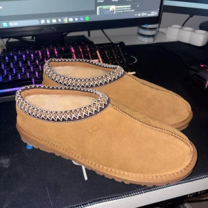 Bruna mocka tofflor från UGG - Säljer ett par supermysiga bruna mocka tofflor från UGG. De har en snygg kantdetalj i mönstrat tyg och är fodrade med mjuk päls för extra värme. Perfekta för att hålla fötterna varma under kalla dagar. Sulan är robust och ger bra grepp. Använda men i mycket bra skick! A kopie 