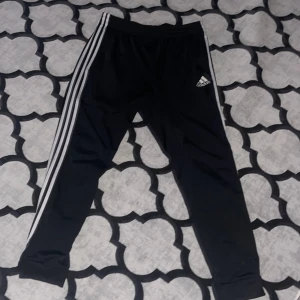 Svarta träningsbyxor från Adidas - Säljer ett par svarta träningsbyxor från Adidas i mycket bra skick. De har de klassiska tre vita ränderna längs sidorna och en elastisk midja för extra komfort. Perfekta för träning eller en avslappnad dag. Adidas-loggan är snyggt placerad på benet.