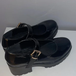 Svarta lackskor från Koi - Snygga svarta lackskor från Koi med en chunky klack och justerbart spänne. Perfekta för en stilren look, oavsett om det är till vardags eller fest. Skorna har en rund tå och en glansig finish.
