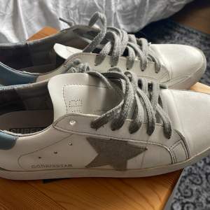 Säljer ett par vita Golden Goose sneakers i bra skick. De har en grå stjärna på sidan och grå snörning. Skorna är perfekta för en avslappnad stil och funkar året runt. Sulan är svart undertill och ger bra grepp. Perfekta för både vardag och fest! Köpt här på plick kan därav inte garantera att dom är autentiska, men dom är fortfarande riktigt feta! Tveka inte att skriva om ni har någon fråga