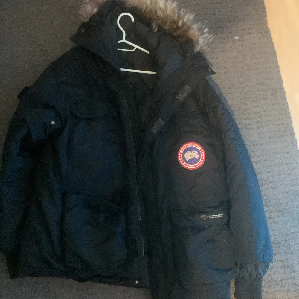 Säljer en svart Canada Goose dunjacka med pälsdetaljer på huvan. Jackan är perfekt för kalla vinterdagar och har en broderad logga på bröstet. Den är i bra skick och har en dragkedja framtill. Supervarm och stilren för vintern! ❄️. Jackor.