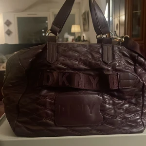 Mörkbrun väska från DKNY - Säljer en snygg mörkbrun quiltad väska från DKNY. Den har ett elegant mönster och är gjord i skinn. Väskan har både handtag och en axelrem för enkel bärning. Insidan är fodrad med rött tyg och har flera fack för bra förvaring. 