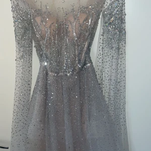 Glittrig långklänning med öppen rygg - Säljer en fantastisk glittrig långklänning i silver med öppen rygg och långa ärmar. Perfekt för nyår eller en glamorös fest! Klänningen har ett vackert fall och är täckt med glittrande paljetter som fångar ljuset. Den är i mycket bra skick och redo att bäras av någon som vill glänsa.