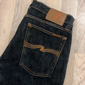 Mörkblå jeans från Nudie Jeans - Säljer ett par snygga mörkblå jeans från Nudie Jeans i mycket bra skick. De har en klassisk femficksdesign med orange sömmar och en bekväm passform. Står strl 30/34 på lapp men är upplagda i längden, längden är mer 32.  Perfekta för både vardag och fest! 🕺 