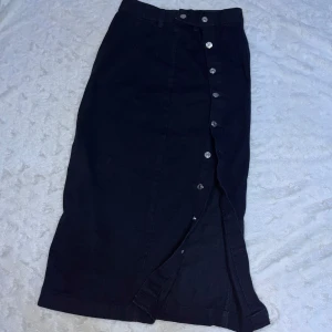 Svart denim långkjol med knappar - Snygg svart denim långkjol med knappar hela vägen ner. Slitsen är framtill och knappar hela vägen ned. Knapparna går inte rakt ned utan har en lugen böj. Storleken är M (38) men passar även bra till storlek S!